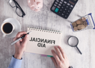 man's hand writing financial aid on notebook with calculator, money and other desk accessories